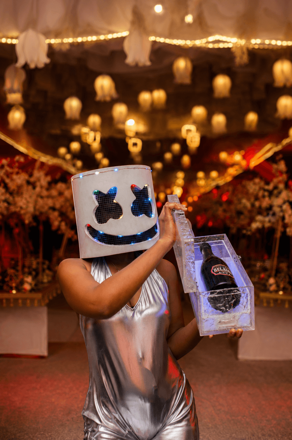 A wide cinematic shot of a luxury garden party at dusk. Guests in elegant evening wear are gathered around a sleek outdoor bar, illuminated by soft golden fairy lights and warm candlelight. The atmosphere is festive yet sophisticated, with the blue hour sky providing a deep contrast to the warm interior glow of the event space. High-end hospitality photography.