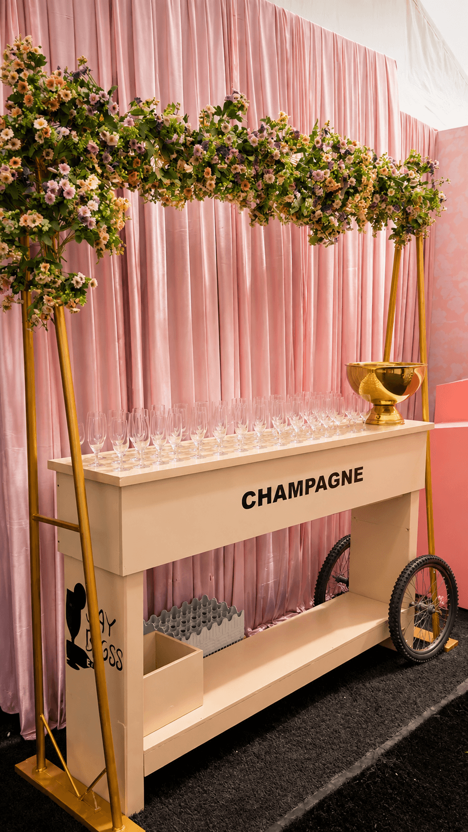 A branded Jaybigss champagne cart trimmed with a floral garland and rows of ready glassware, set against a draped pink backdrop